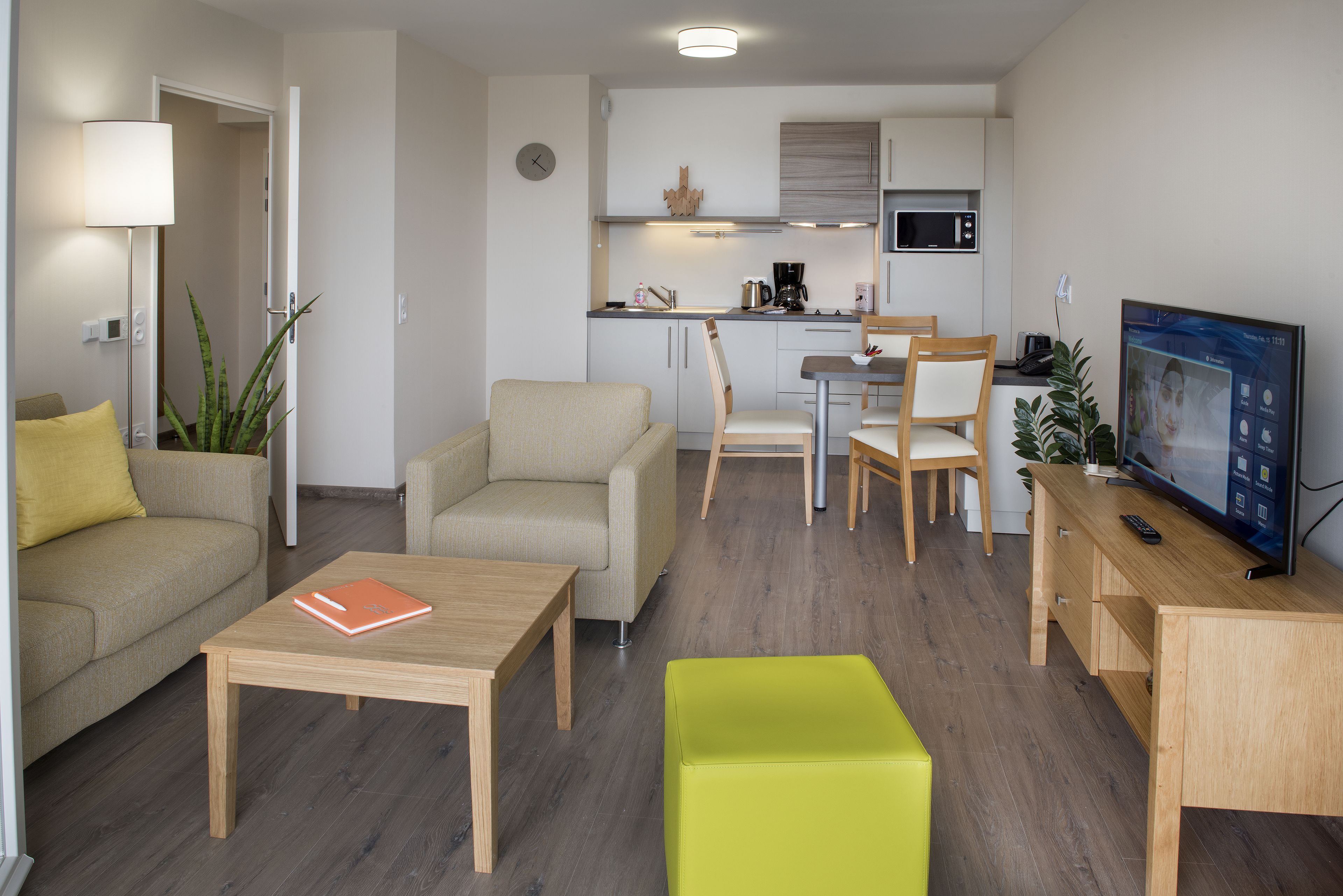 A modern living room with a sofa, coffee table, and a TV. There is a kitchen area with a microwave and a dining table.