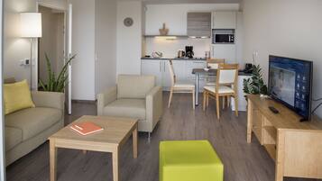 A modern living room with a sofa, coffee table, and a TV. There is a kitchen area with a microwave and a dining table.