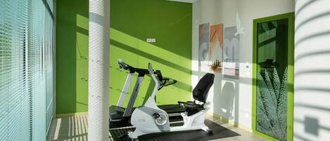 A fitness room with treadmills and a stationary bike, green walls, and a large window with blinds.