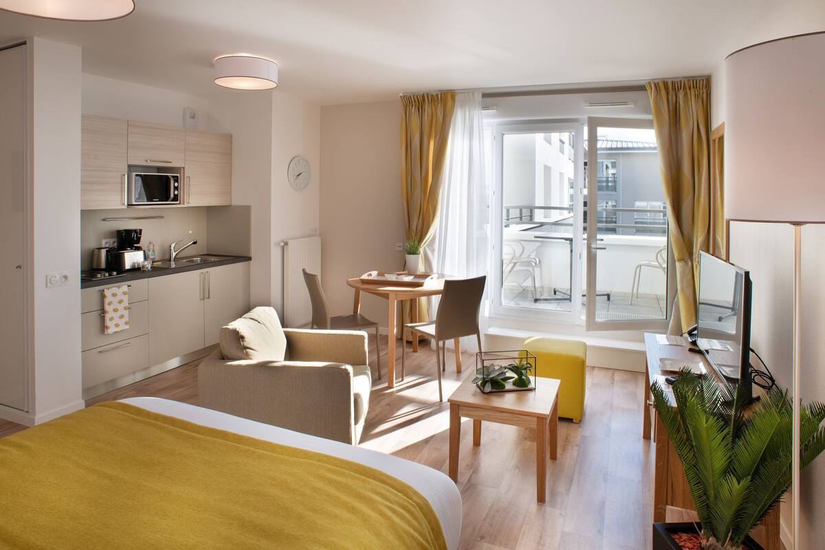 A modern hotel room with a bed, a desk with a computer, a dining area, a kitchen, and a balcony with curtains.