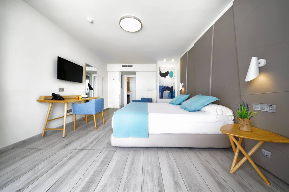 Habitación de hotel moderna con una cama grande, un escritorio con silla, un televisor montado en la pared y una mesita de noche con una planta.
