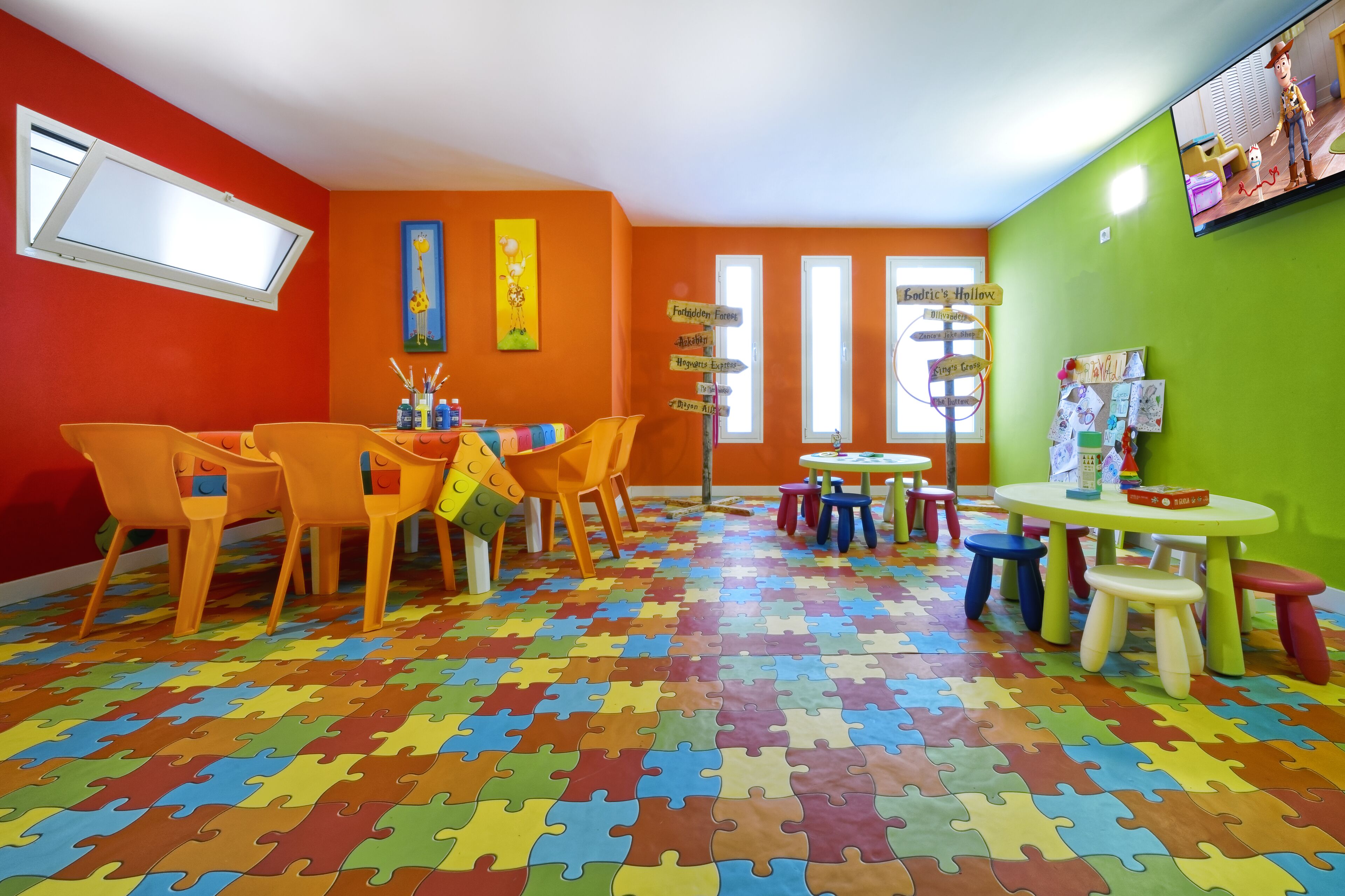 Une salle de jeux aux couleurs vives, avec un sol décoré de motifs de puzzle, des murs orange et un téléviseur fixé au mur vert.