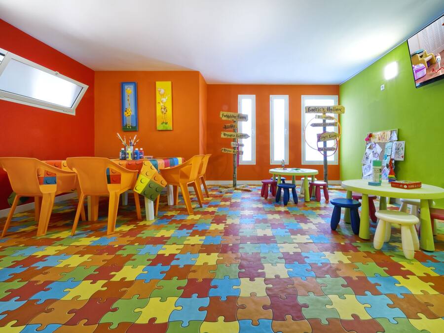 Uma sala de jogos vibrante, com piso estampado como quebra-cabeça, paredes laranja e uma televisão fixada na parede verde.