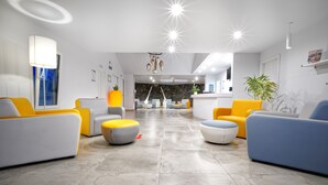 Un moderno hall de hotel con mostrador de recepción, zona de estar con sofás amarillos y blancos, y un fondo de pared de piedra.