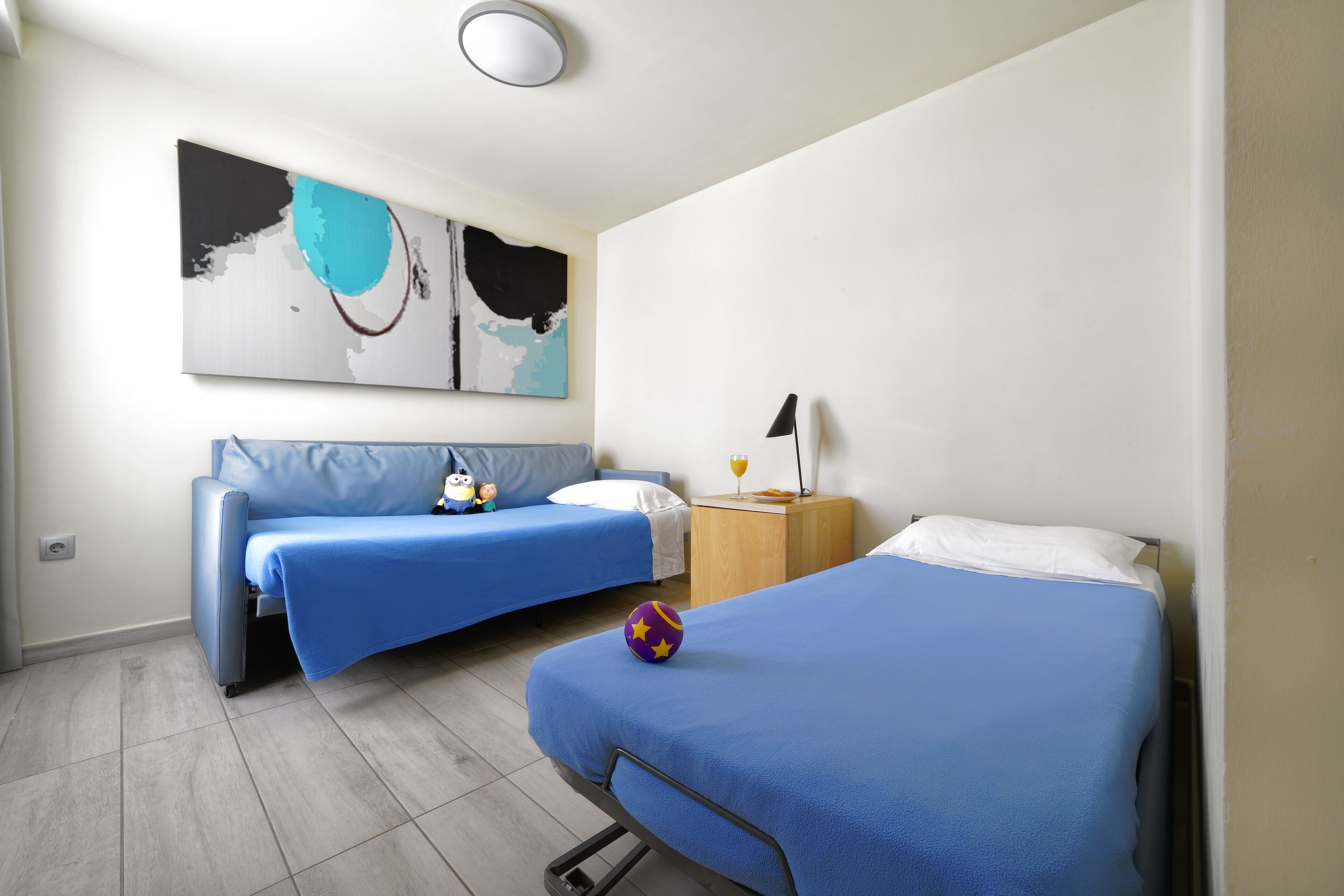 A room with two beds, a sofa, a wall art piece, and a nightstand with a lamp.