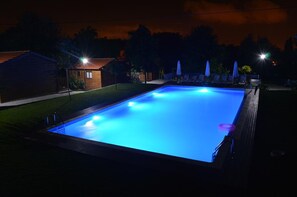 Una piscina rectangular con luz azul durante la noche, rodeada por una terraza de madera y un cobertizo pequeño.