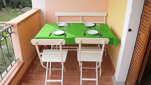 Un pequeño balcón exterior con una mesa para dos personas, sillas blancas y un mantel verde.