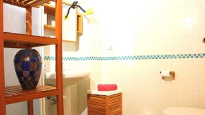 Baño con lavamanos, inodoro y un pequeño taburete de madera con una toalla roja. Se observa una estantería con un florero decorativo y un espejo con una toalla colgando.