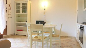 Una cocina pequeña y bien iluminada con una mesa y sillas, un armario blanco con puertas de vidrio, y una lámpara fijada a la pared.