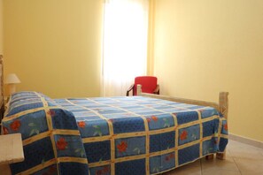 A room with a bed covered in a floral-patterned bedspread, a red chair, and a window with curtains. - Residenza del Comandante (Santa Teresa di Gallura)