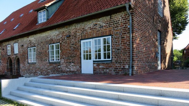 A brick building with a red-tiled roof, white-framed windows, and a set of concrete steps leading to a door. - Gästehaus Klein Grönau (Lübeck)