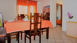 A dining area with a table set for four, wooden chairs, a window with orange curtains, and a painting on the wall. - Residence Ostro (Santa Teresa di Gallura)