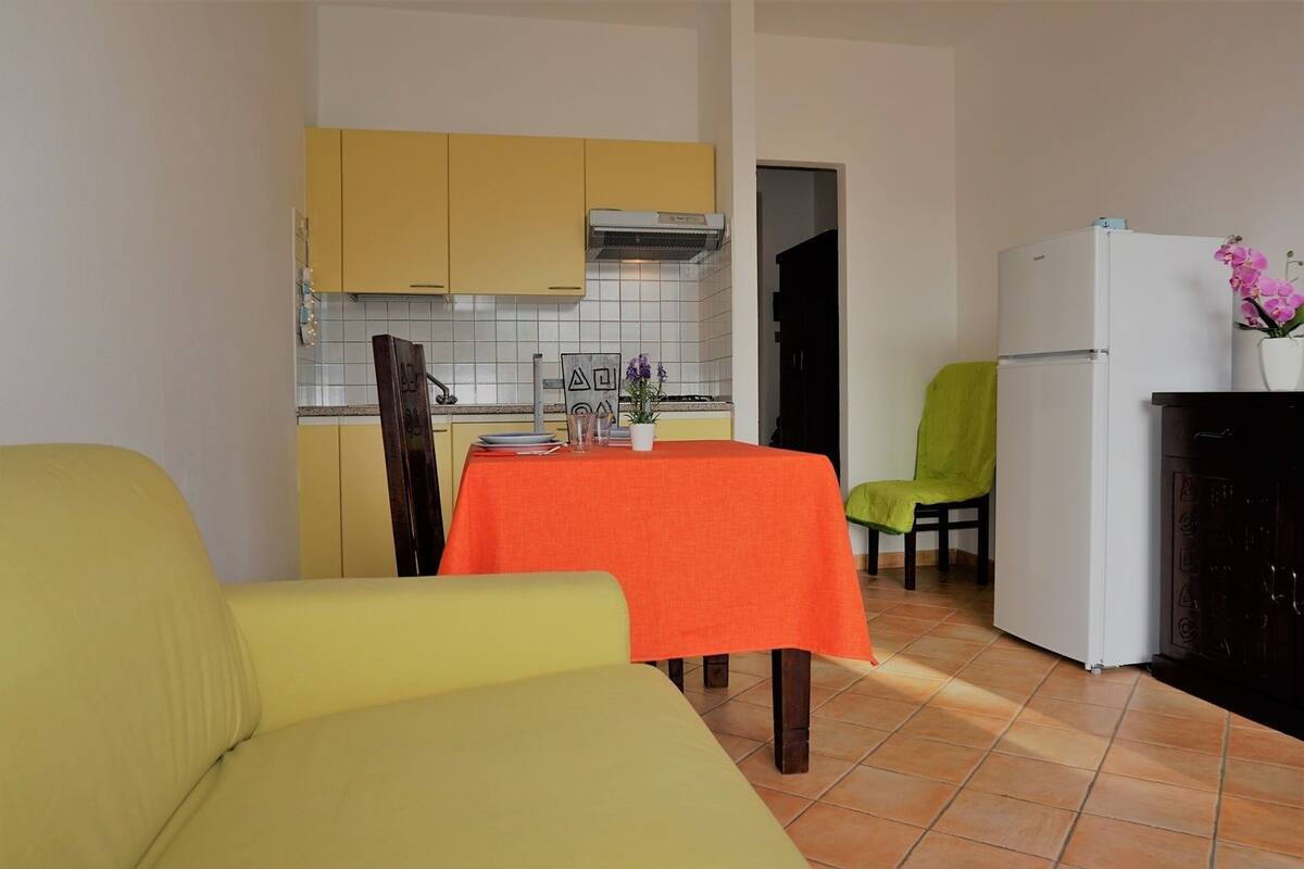 Une cuisine de taille modeste, avec des meubles de couleur jaune, un réfrigérateur blanc et une table à manger recouverte d’une nappe orange.