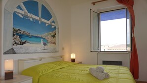 A bedroom with a bed, a nightstand, a lamp, a window with curtains, and a painting of a beach scene. - Residence Levante (Santa Teresa di Gallura)