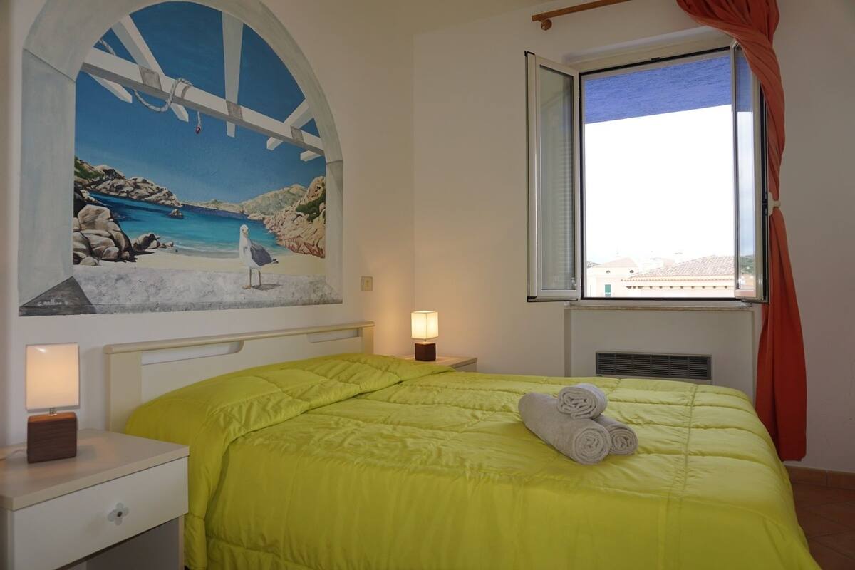 Une chambre à coucher avec un lit, une table de chevet, une lampe, une fenêtre avec des rideaux et un tableau représentant une scène de plage.