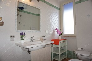 Une salle de bain avec un lavabo blanc, un miroir et des toilettes. Une fenêtre laisse entrer la lumière naturelle et une étagère verte est ornée d’un vase de fleurs.