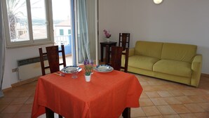 A dining area with a table set for two, a yellow sofa, and a view of the outdoors through large windows. - Residence Levante (Santa Teresa di Gallura)