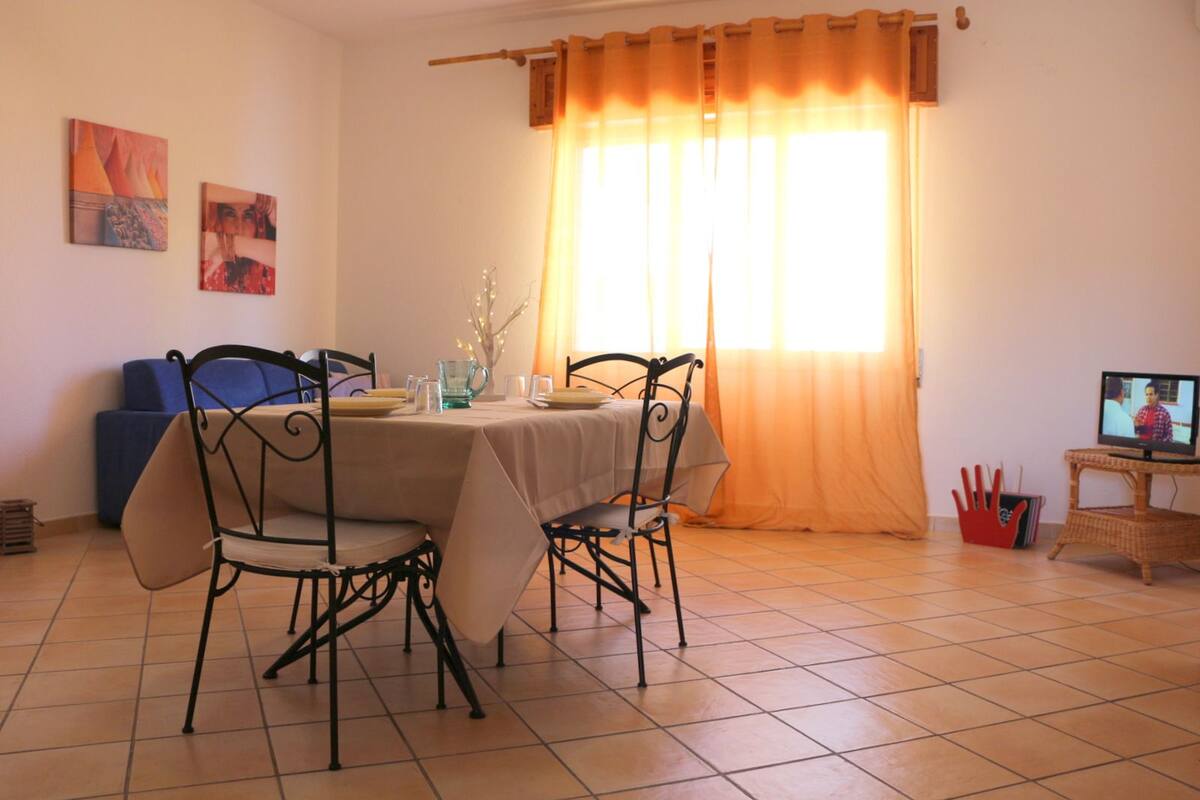 A dining area with a table set for four, chairs, a television on a stand, and a window with curtains.