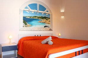 Quarto com cama, mesa de cabeceira, abajur e pintura de uma paisagem costeira.