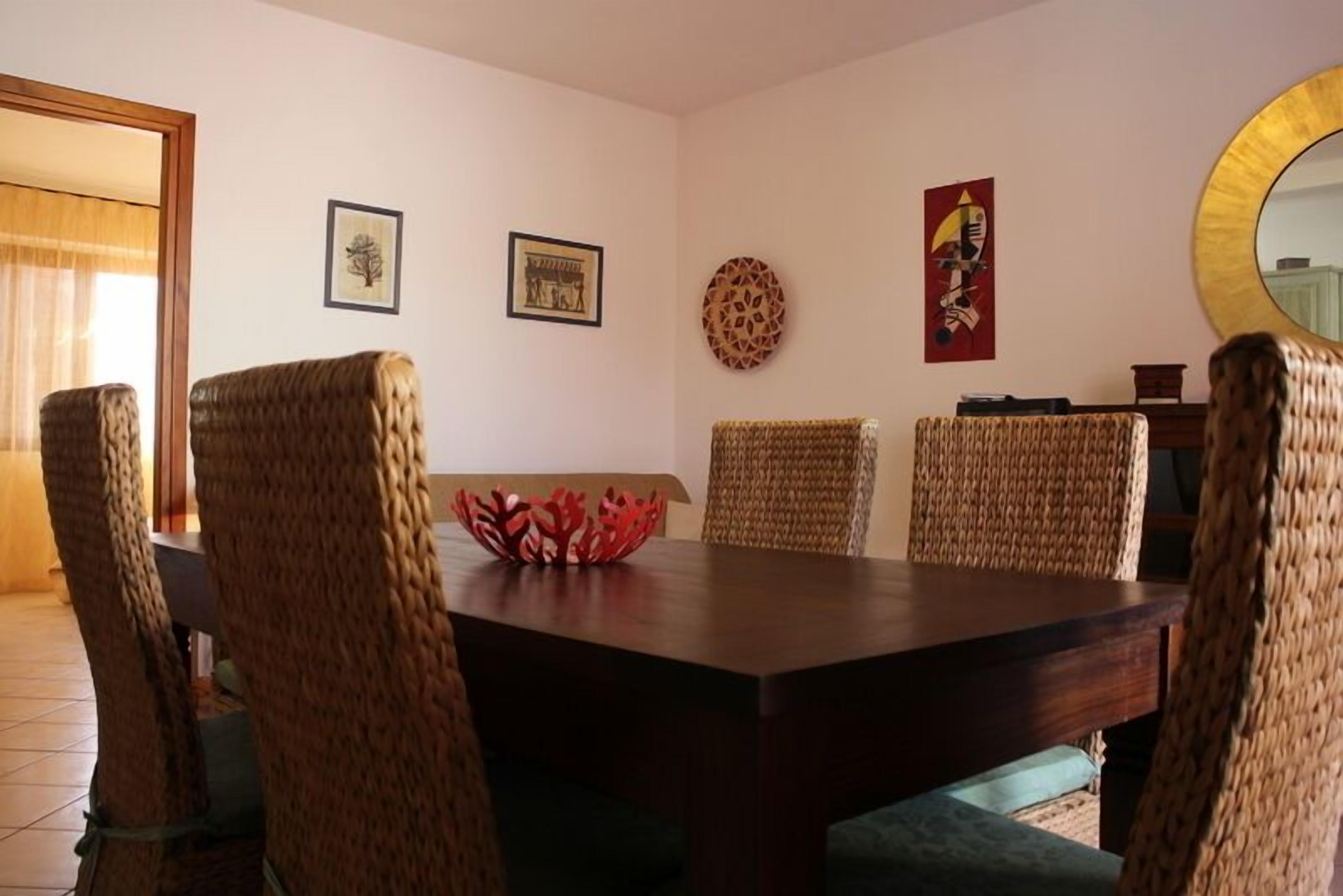 A dining area with a dark wooden table and woven chairs. There are framed artworks on the walls and a decorative bowl on the table.