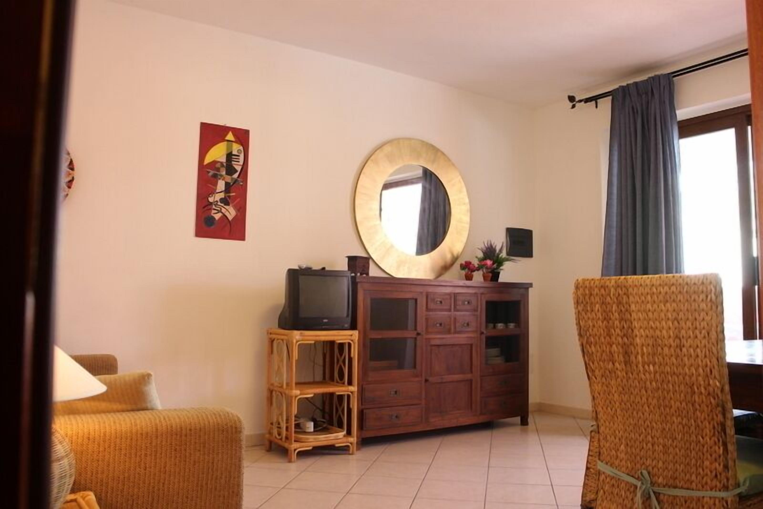 Un salon avec un canapé, un téléviseur posé sur un meuble en bois, un miroir rond et un fauteuil en osier.
