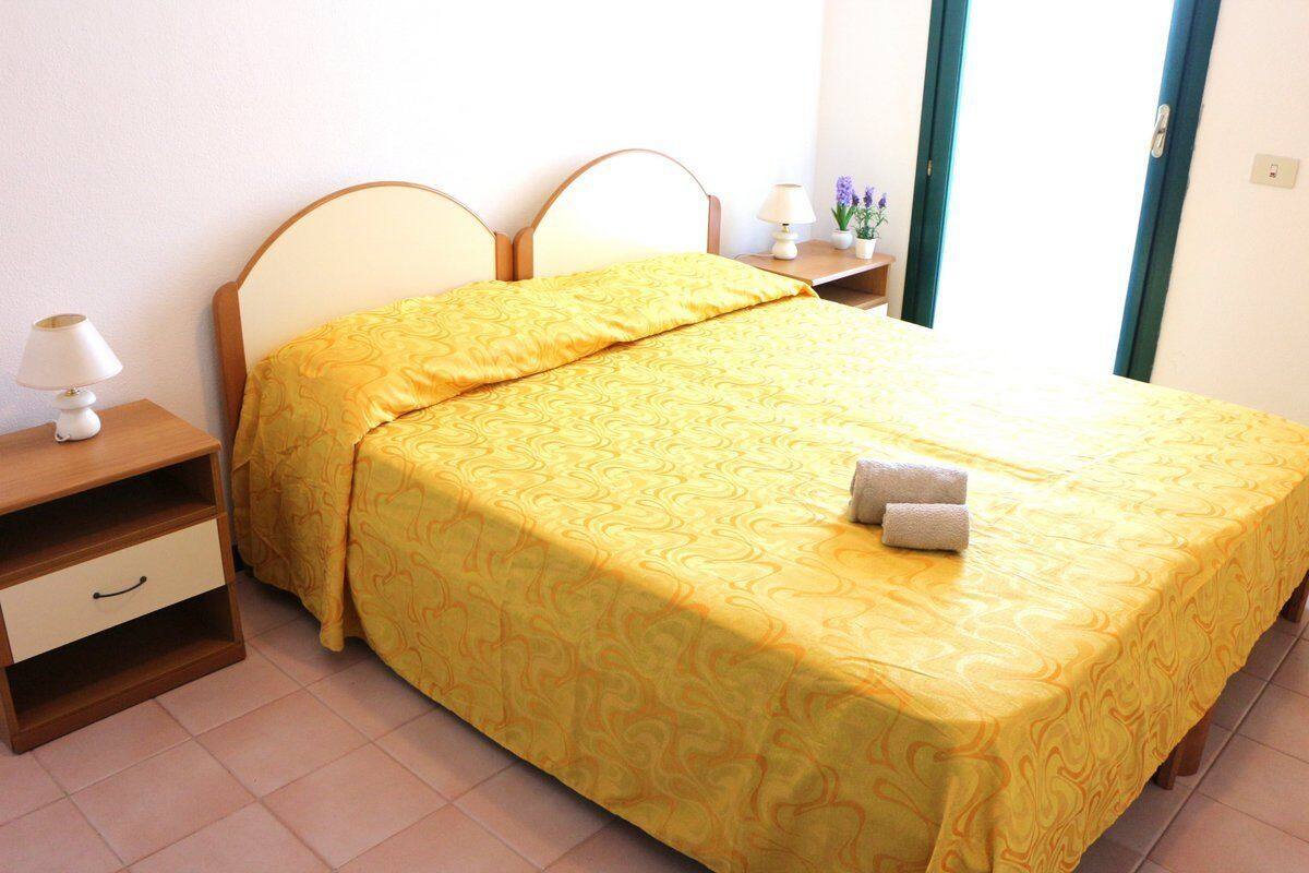 A neatly made bed with a yellow patterned bedspread and pillows, a bedside table with a lamp, and a small nightstand with a vase of flowers.