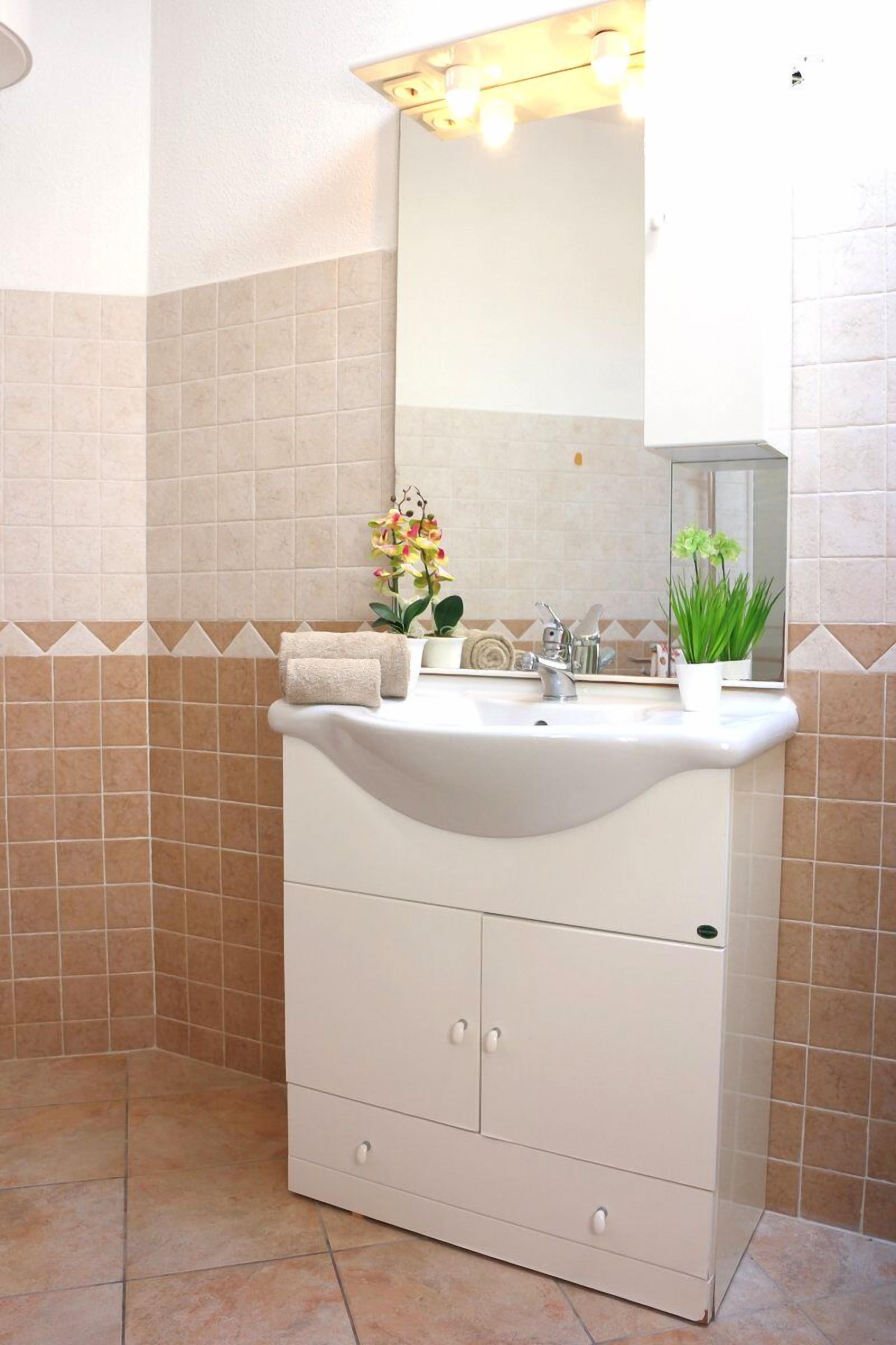 A bathroom with a white sink, mirror, and cabinet, beige tiles, and a potted plant.