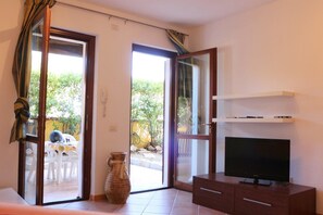 Ein Wohnzimmer mit Fernseher auf einer Holzkommode, einer Vase und einer Glastür, die zu einem Außenbereich mit Tisch und Stühlen führt.
