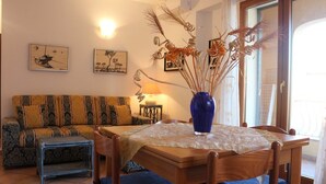 A cozy living room with a sofa, a dining table with chairs, a blue vase with dried plants, and artwork on the walls. - Rena Bianca Apartment (Santa Teresa di Gallura)