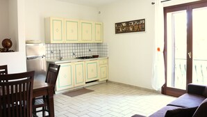 A compact kitchen with white cabinets, a dining table with chairs, and a sofa in a bright living area with a view through a sliding door. - Casette del Centro (Santa Teresa di Gallura)