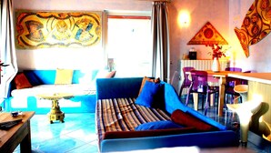 A living room with a sofa, a dining table, and chairs. There is a window with curtains, a wall with a tapestry, and a lamp on the table. - Rena Bianca Beach House (Santa Teresa di Gallura)
