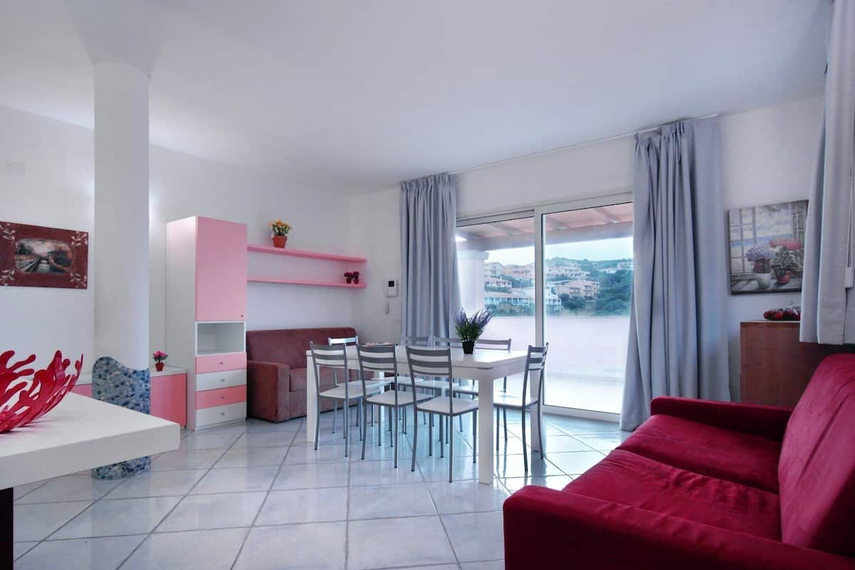 Un salon moderne avec un canapé rouge, une table à manger avec des chaises, un meuble rose et une grande fenêtre donnant sur des immeubles.