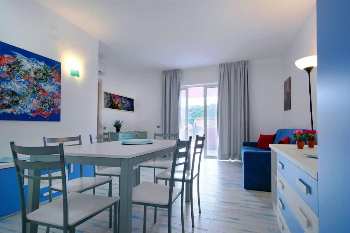 Un salon moderne avec un coin repas, comprenant un canapé bleu, une table à manger avec des chaises et un grand tableau abstrait accroché au mur.