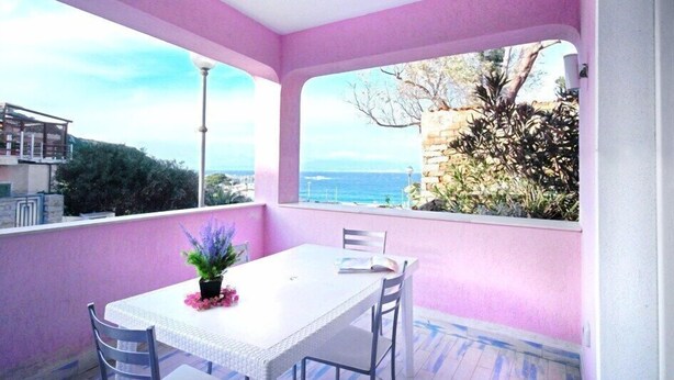 A balcony with a white table and chairs, a potted plant, and a view of the sea and buildings. - Rena Bianca Beach House (Santa Teresa di Gallura)