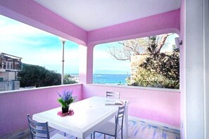 A balcony with a white table and chairs, a potted plant, and a view of the sea and buildings.