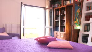 A bedroom with a purple bed, a wooden bookshelf, and a window with a view. - Casa Seby (Santa Teresa di Gallura)