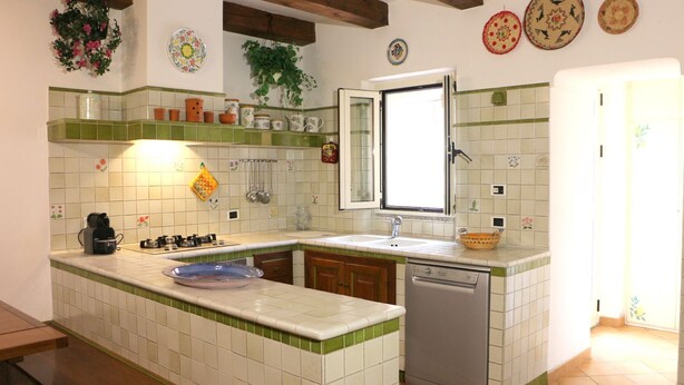A kitchen with a green and white tiled backsplash, a built-in dishwasher, and a window above the sink. - Casa Seby (Santa Teresa di Gallura)