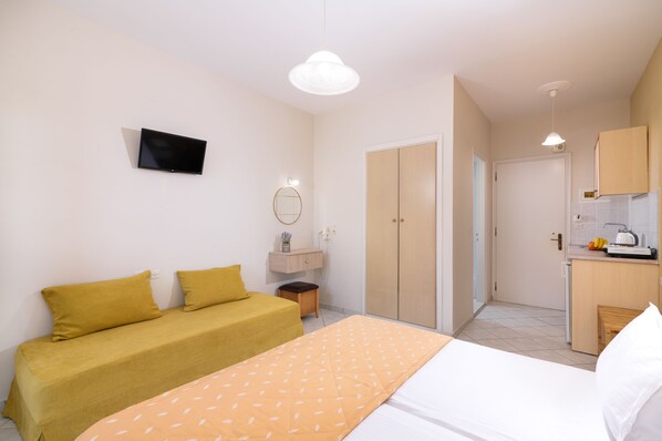 A bedroom with a bed, a sofa, a TV, a wardrobe, a small table, and a kitchen area.