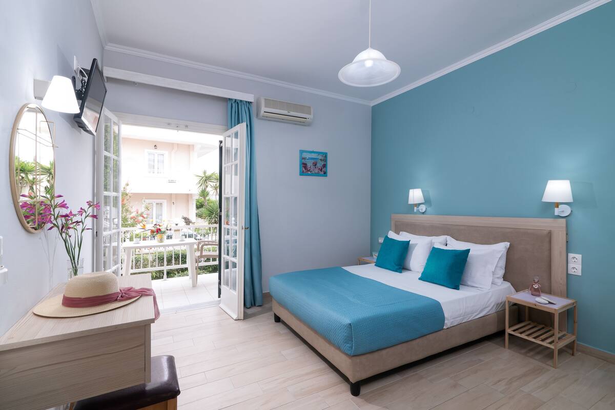 Un dormitorio con cama, mesitas de noche, escritorio, silla, lámpara colgante y vista al balcón exterior.