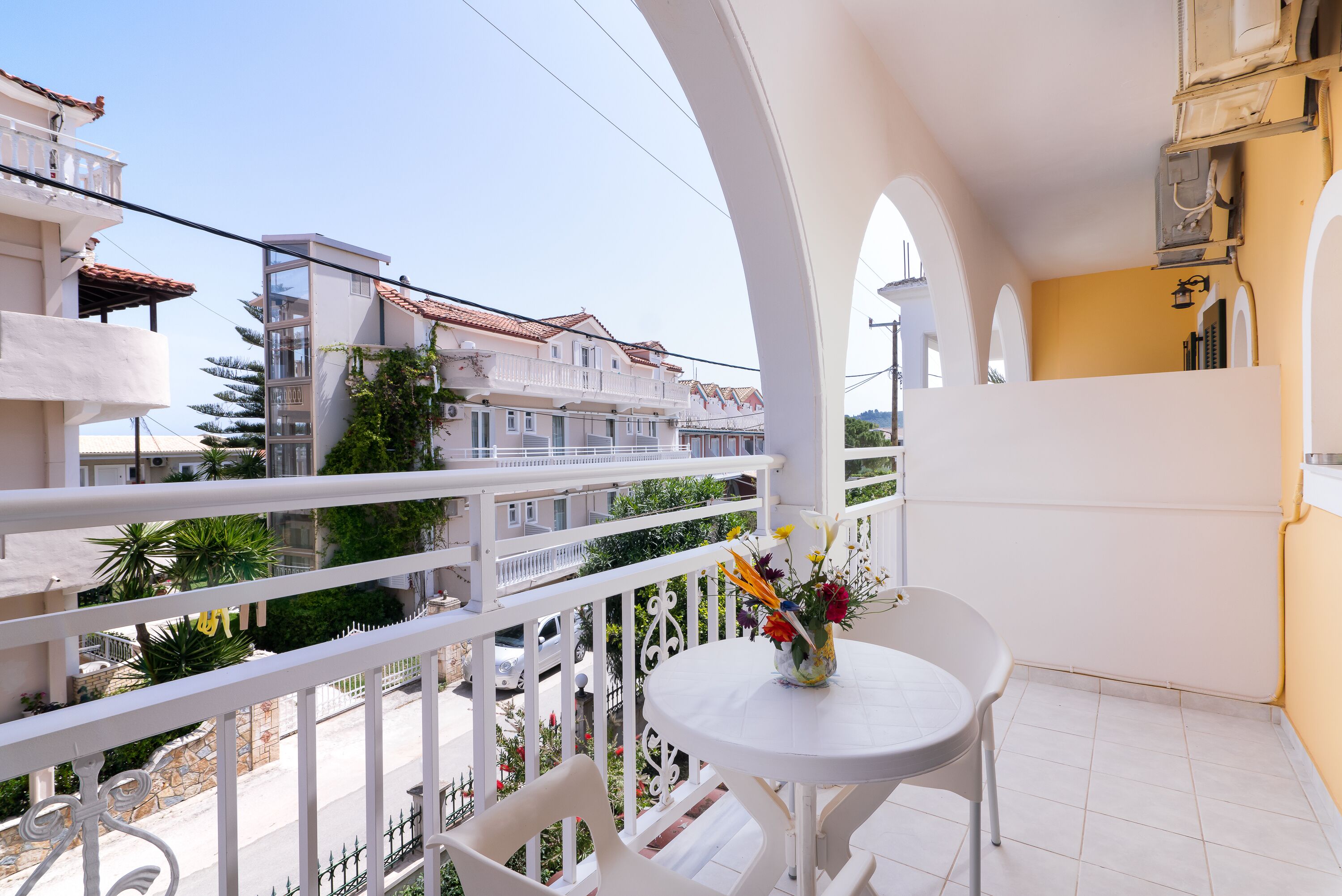 Un balcón con una mesa y sillas blancas, un jarrón de flores y vistas a edificios vecinos.