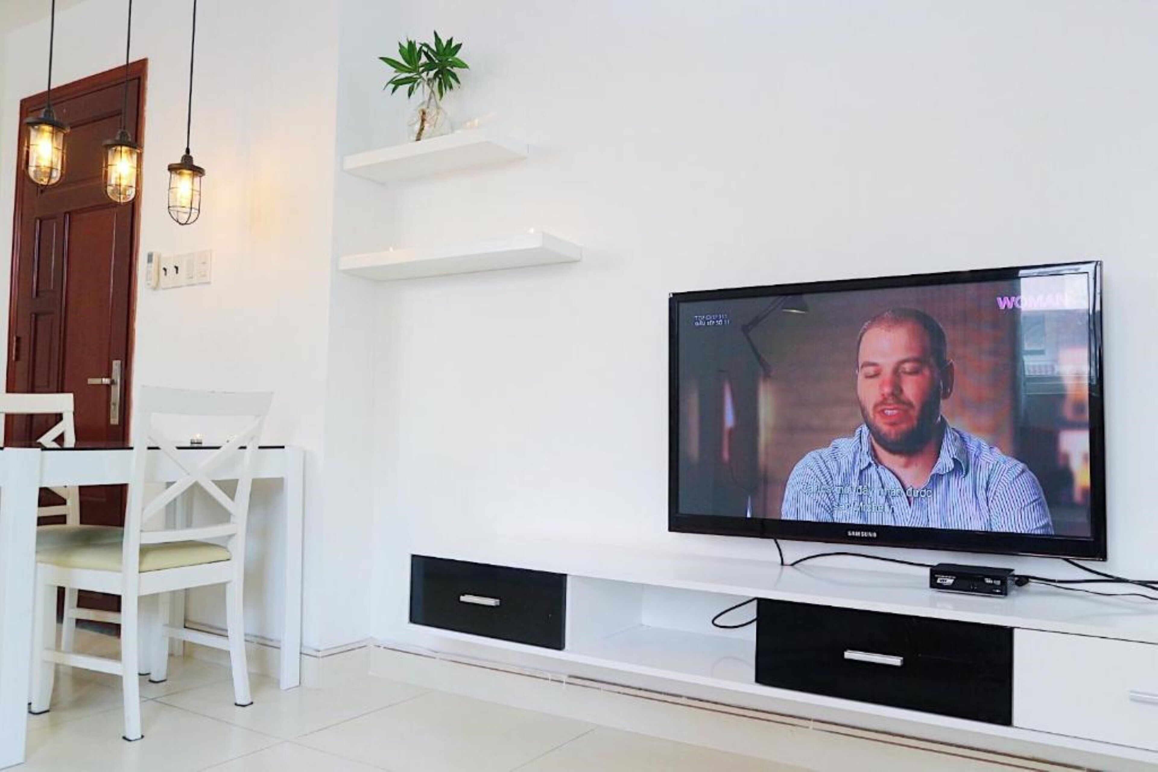 Una sala de estar moderna con un televisor de pantalla plana montado en un mueble blanco, una puerta de madera y una mesa de comedor blanca con sillas.