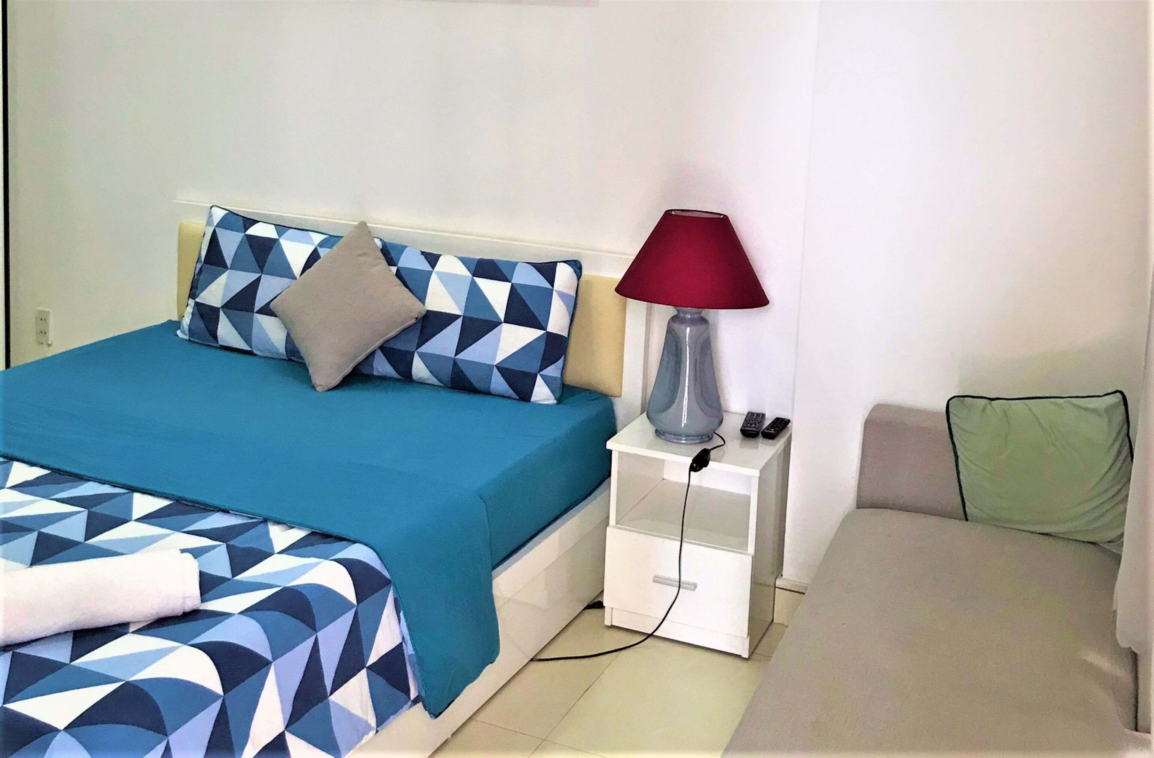 Una cama con una colcha azul y blanca estampada, una lámpara de noche roja y una mesita de noche blanca con un control remoto.
