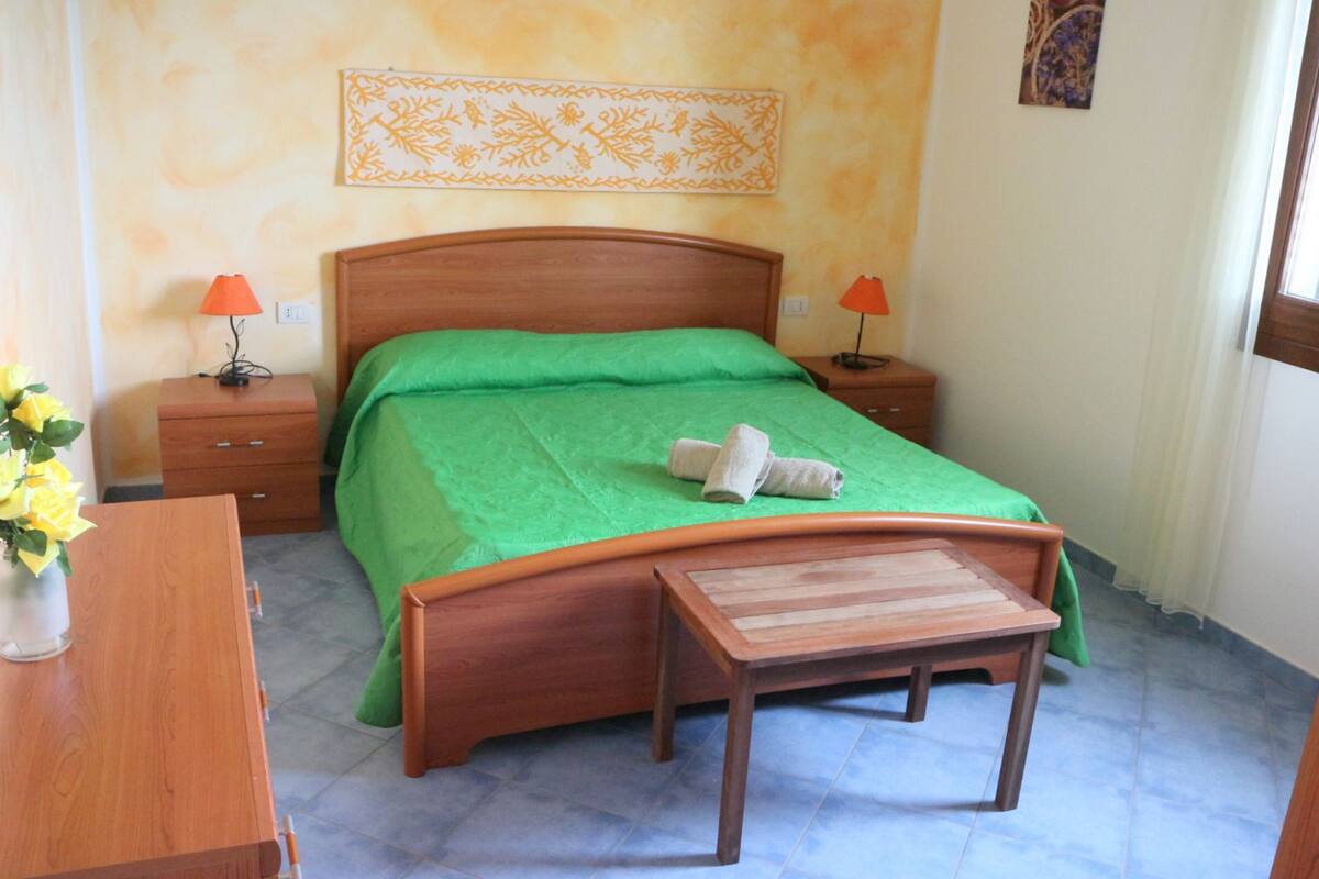A bedroom with a wooden bed, green bedspread, two bedside tables with lamps, a wooden nightstand, and a small wooden bench.