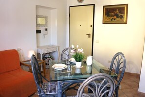 Een eethoek met een glazen tafel, stoelen en een vaas bloemen. Achtergrond is een deur en aan de muur een schilderij.
