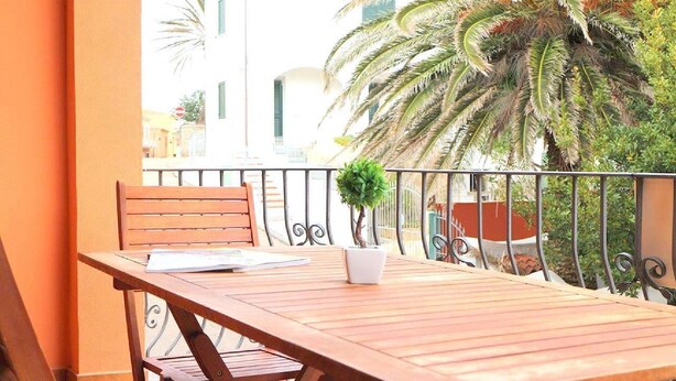A wooden table and chair on a balcony with a railing, a small potted plant, and a view of buildings and trees. - Gemelle di Cairoli (Santa Teresa di Gallura)