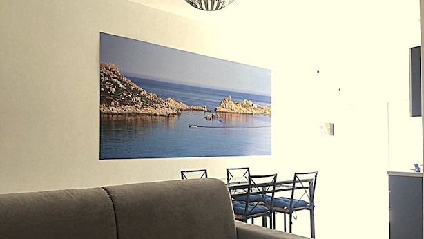 A living room with a grey sofa, a dining table with chairs, and a large painting of a coastal landscape on the wall. - Tiffany House (Santa Teresa di Gallura)