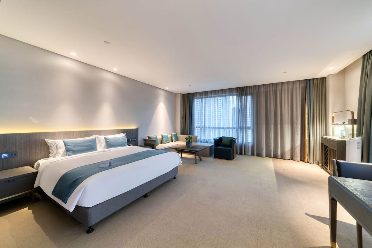 A modern hotel room with a large bed, a sofa, a desk, and a view of the city through the window.