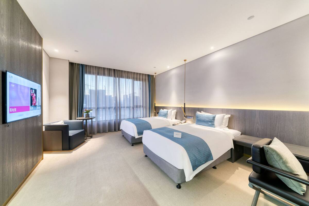 A modern hotel room with two beds, a TV, a desk, and a large window with curtains.