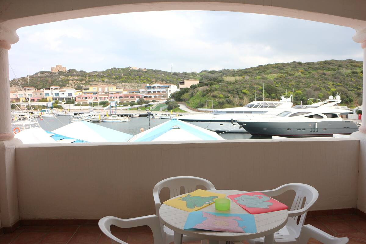 A balcony with a table and chairs, overlooking a marina with yachts and a hillside town.