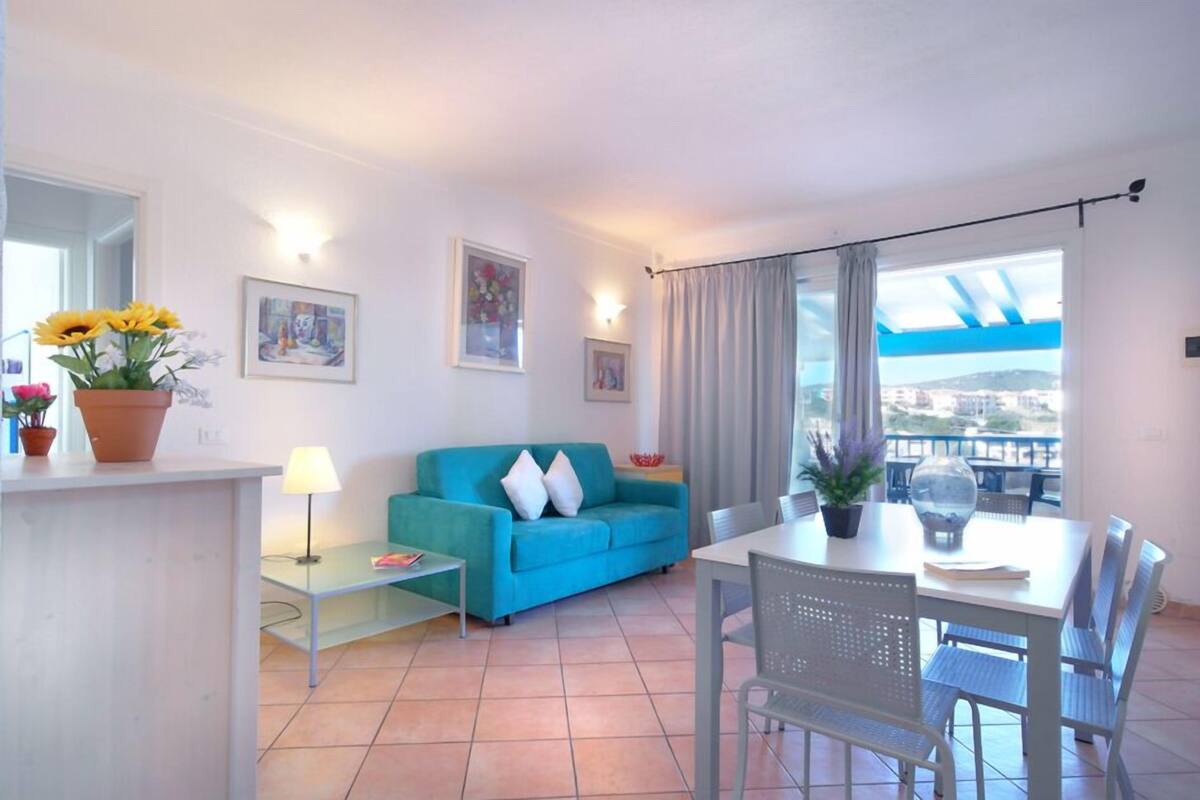 A living room with a sofa, a dining table with chairs, a small table with a lamp, and a large window with a view.